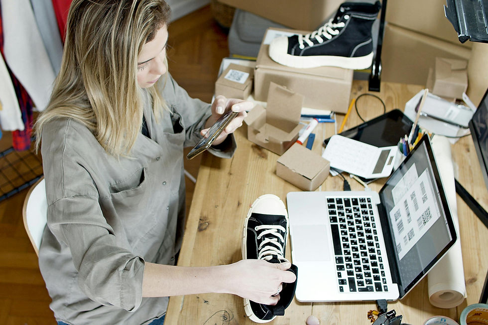 A woman working on her laptop while taking a picture of a shoe with her smartphone, representing an online store owner creating product content for e-commerce marketing.