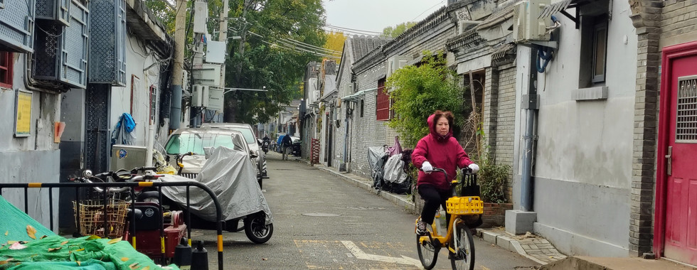 Moradora pedalando bicicleta compartilhada amarela da Meituan no Lishi Hutong entre casas de tijolo cinza e portões vermelhos, Dongcheng, Pequim