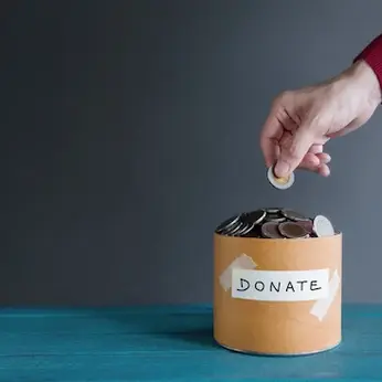 Hand placing a coin into a tzedakah box, emphasizing the daily act of giving as a mitzvah.