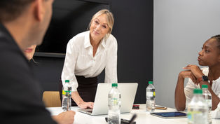 Smiling woman in a white blouse leads a meeting around a laptop, with colleagues listening at a conference table.