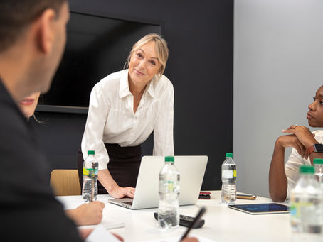 Smiling woman in a white blouse leads a meeting around a laptop, with colleagues listening at a conference table.