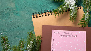 Notepad with "New Year's Resolutions" on pink paper, blank list, surrounded by snow-dusted pine branches on a green textured surface.