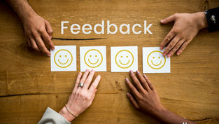 Hands on a wooden table arranging smiley cards under the word "Feedback."