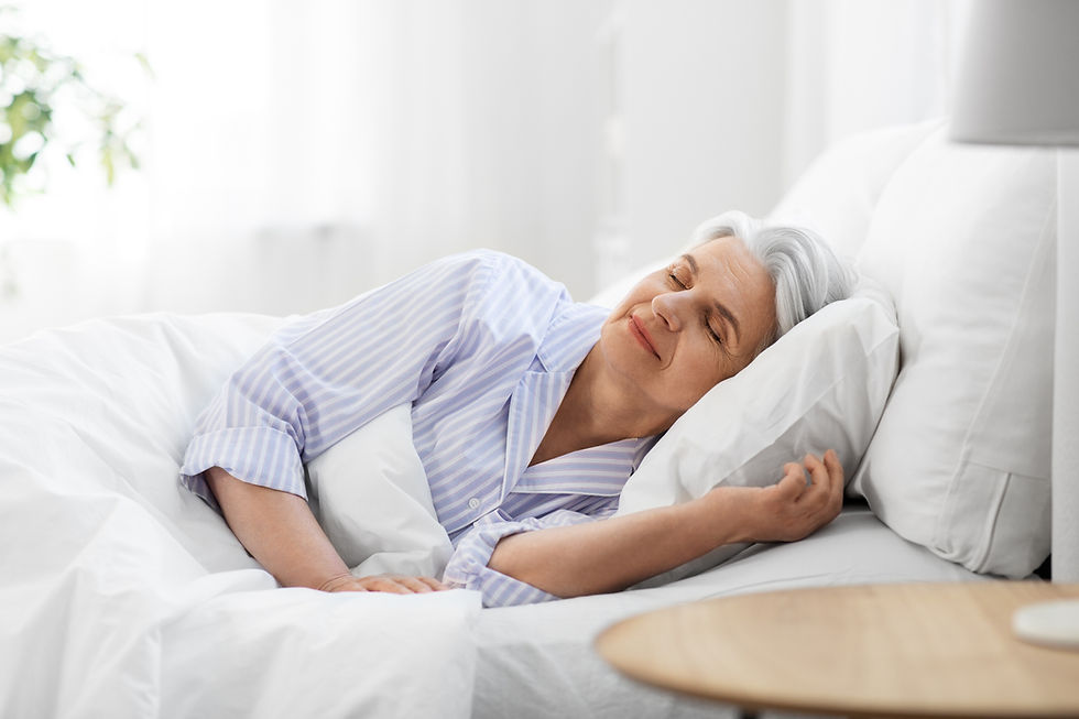 An older woman with gray hair sleeps peacefully in a bright bedroom. She's wearing a striped pajama top, lying on white bedding.