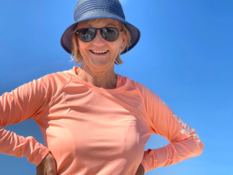 Smiling woman in blue sunhat and sunglasses wears a peach long-sleeve top against a clear blue sky.