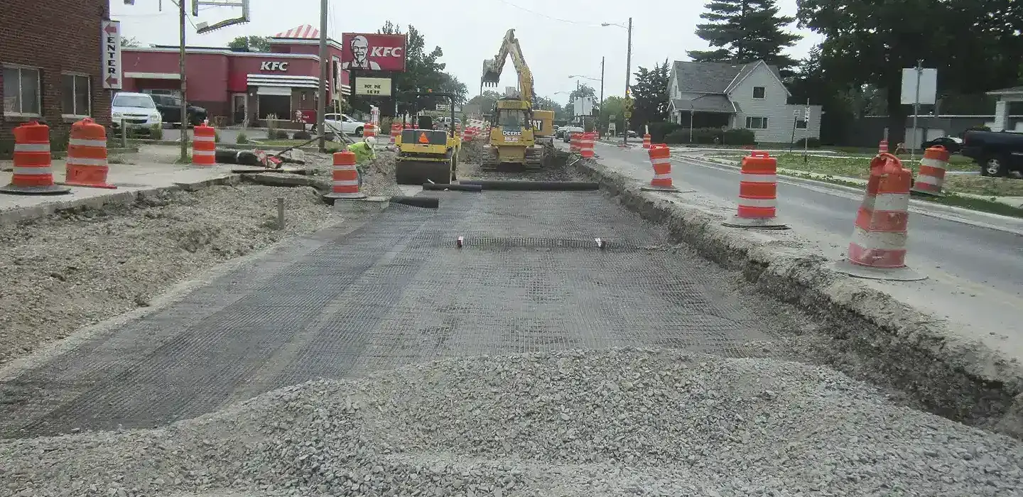 Active construction site of a full depth asphalt roadway reclamation project featuring a large pile of gravel, safety barrels, and heavy machinery.