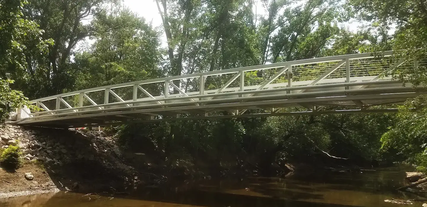 Replacement of a deficient pedestrian bridge at the University of Toledo. The bridge is running over a river. There are trees in the background.