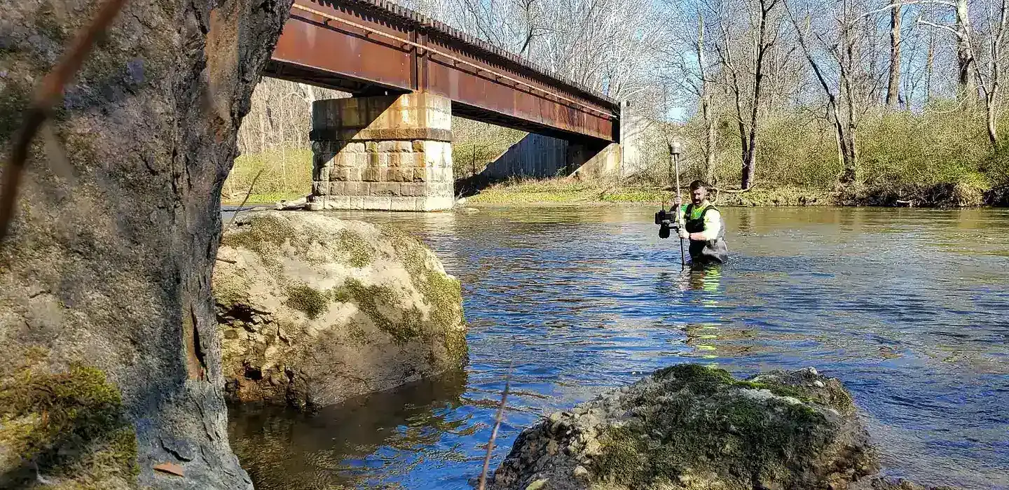 surveyor wading through a river with a GPS unit with rocks a bridge and trees surrounding