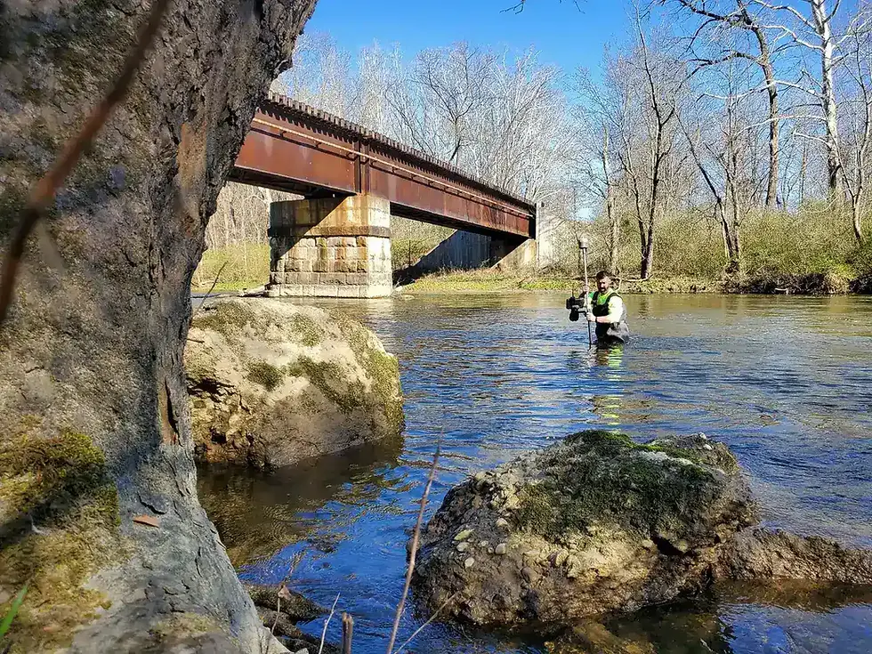 surveyor wading through a river with a GPS unit with rocks a bridge and trees surrounding