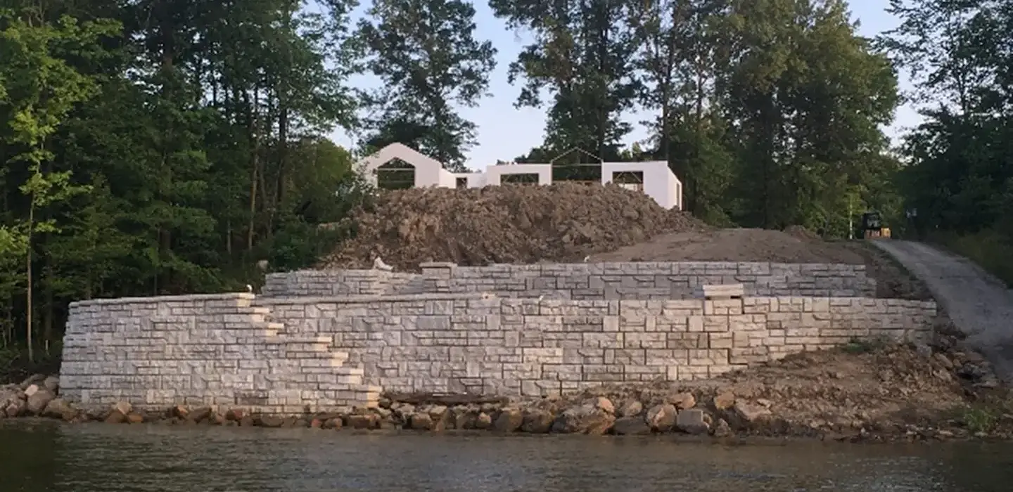 Retaining wall made of concrete bricks. There is a lake on one side of the retaining wall and there is a house and dirt on an incline on the other side. 