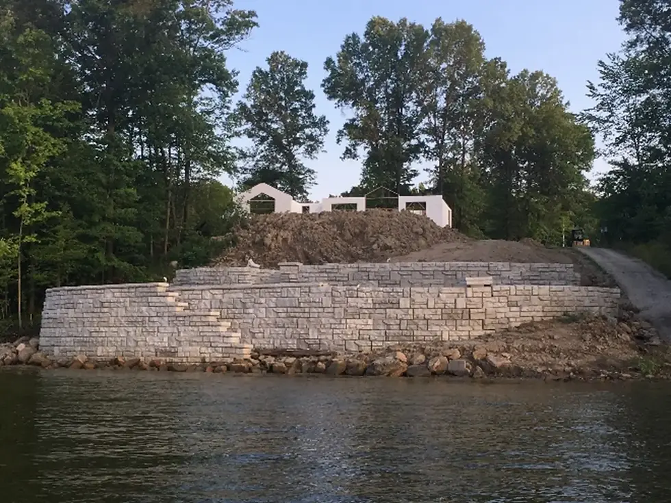 Retaining wall made of concrete bricks. There is a lake on one side of the retaining wall and there is a house and dirt on an incline on the other side. 