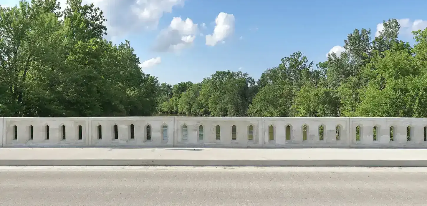Concrete railing bridge feature and pedestrian walkway adjacent to the grooved concrete deck surface. Trees and river in the background.