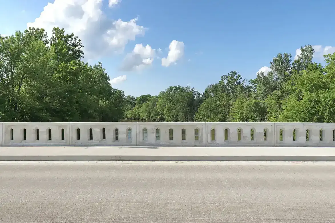 Concrete railing bridge feature and pedestrian walkway adjacent to the grooved concrete deck surface. Trees and river in the background.
