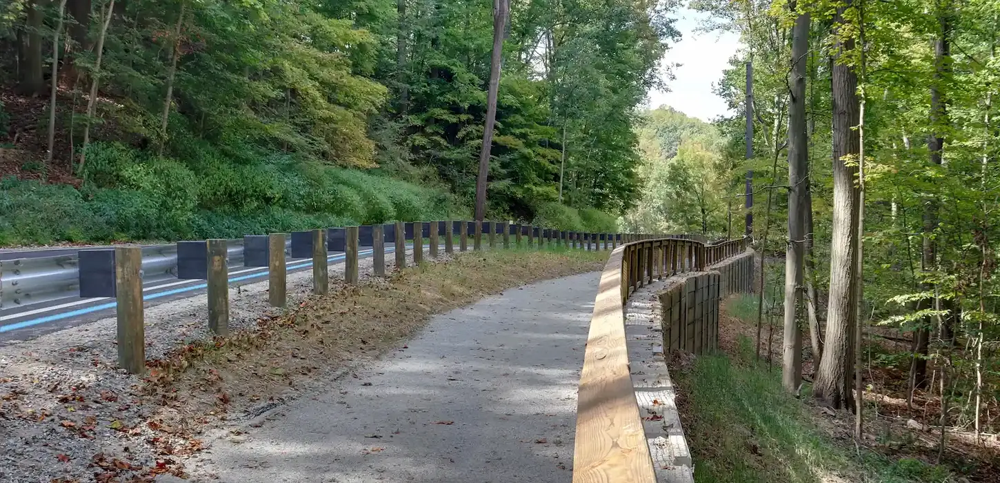 Retaining wall with MUP trail running through metro park. There are trees in the background and the trail is fenced in running parallel to the main road.