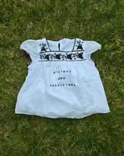 A light, white cap-sleeve shirt with a flared bottom and black machine-embroidered flowers around the neck. It has "Vicious and Suspicious" printed across it in black lettering.