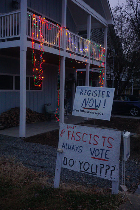 A sign in someone's yard that says "Fascists always vote. Do you???" with a smaller sign that says "Register Now!" above it. Both are on white-painted boards of some kind. Christmas lights are strung along an upper balcony in the background.