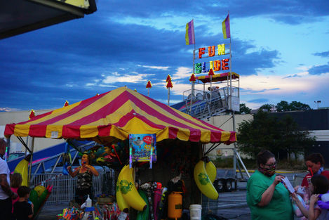 A scene from a parking lot carnival with a tall slide rising in the background. It has colorful lighted letters that say "FUN SLIDE" with flags on either side. The foreground has a red and yellow striped game tent with large stuffed bananas hanging off the side. There are several carnival-goers around it. The sky is light blue with dark clouds.