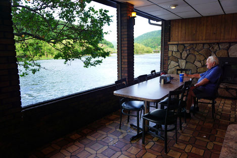 An elderly man in a blue shirt sitting alone at a long table looking out at the Greenbrier River through large windows. The wall behind him is fake stone and wood paneling. The floor is a wood-print geometric linoleum.