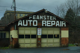 A large brown garage with two tan garage doors and a sign that says "FEAMSTER AUTO REPAIR" in large 1950's-style white letters. The lettering is within the peak of the garage with "FEAMSTER" slightly smaller than "AUTO REPAIR." It is a dark and cloudy day.