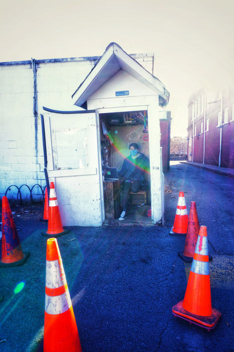 A masked parking attendant sitting in a tiny white shack within a circle of orange traffic cones. The shack is a converted bathroom and still says "Restroom" in white letters on a tiny black sign in the peak. The image is oversaturated so that the cones are very bright, and the pavement looks dark blue. There is a semicircular rainbow sun flare across the center of the image (and the attendant's face).