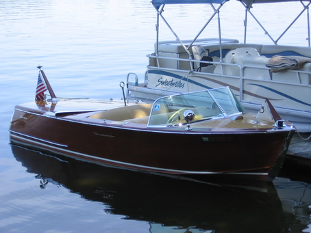 A beautifully restored vintage wooden runabout boat with a rich mahogany finish is docked on calm water beside a modern pontoon boat. The classic boat features a polished wood deck, chrome accents, a wraparound windshield, cream-colored seats, and an American flag at the stern, showcasing timeless craftsmanship and elegant boat seating design.