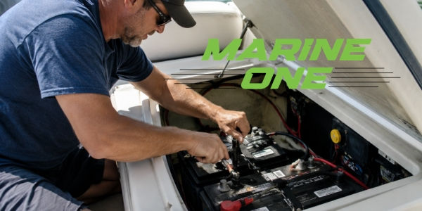 Man in sunglasses works on boat batteries under a hatch. Text "MARINE ONE" in green. Bright daylight setting.