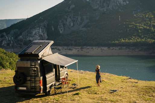 Camper van with a rooftop tent and extended awning parked beside a scenic mountain lake, while a person in a sun hat enjoys the peaceful waterfront view. Ideal image representing RV travel, off-grid camping, outdoor adventure, and exploring nature.