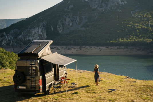 Camper van with a rooftop tent and extended awning parked beside a scenic mountain lake, while a person in a sun hat enjoys the peaceful waterfront view. Ideal image representing RV travel, off-grid camping, outdoor adventure, and exploring nature.
