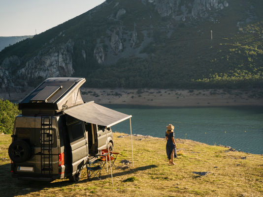 Camper van with a rooftop tent and extended awning parked beside a scenic mountain lake, while a person in a sun hat enjoys the peaceful waterfront view. Ideal image representing RV travel, off-grid camping, outdoor adventure, and exploring nature.