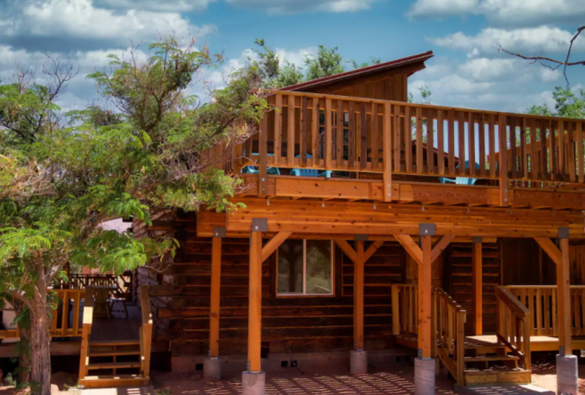 Exterior view of the upper deck of Cliff View Cabin vacation rental, surrounded by lush trees under a beautiful blue sky with puffy white clouds
