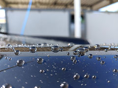 A close-up of a car’s surface with water droplets beading on a ceramic-glass coating, showcasing the hydrophobic properties and high-gloss finish achieved through Detail Koning’s expert paint protection. Een close-up van het oppervlak van een auto met waterdruppels die op een keramische glascoating parelen, wat de hydrofobe eigenschappen en de hoogglanzende afwerking laat zien die wordt bereikt door de deskundige lakbescherming van Detail Koning.