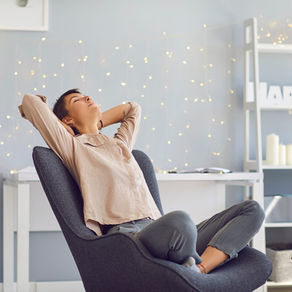 Woman in a gray chair, relaxed with hands behind head. Soft lighting with fairy lights in the background, creating a calm atmosphere.