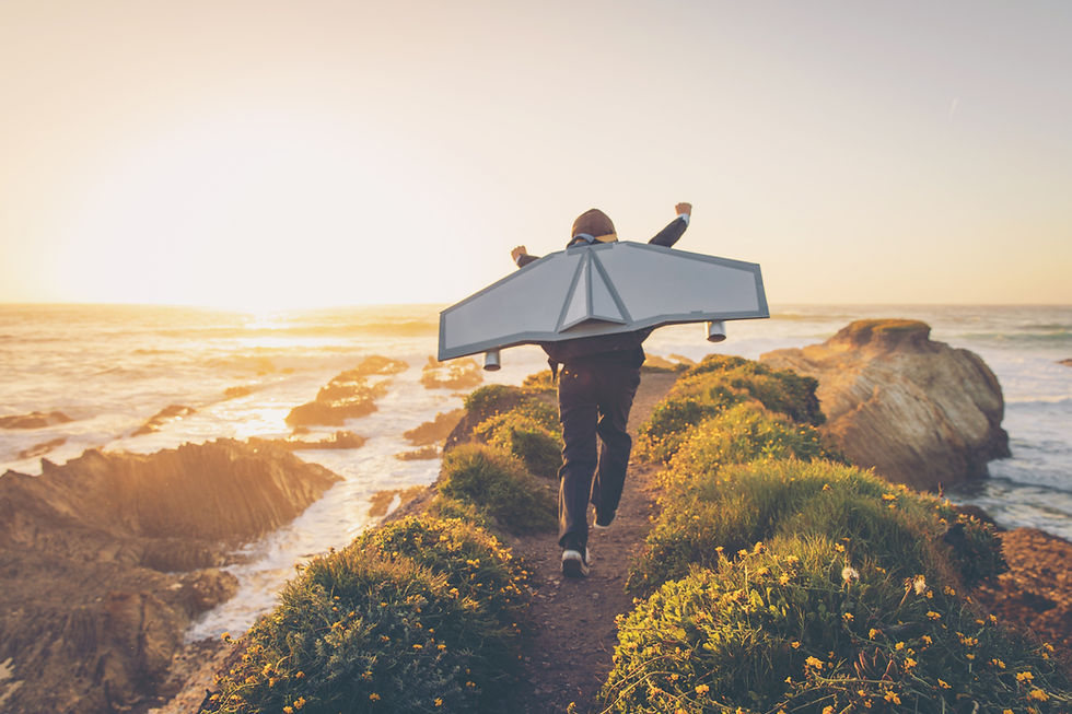 Child with cardboard wings runs on a grassy cliff path at sunset, overlooking a rocky ocean coast, evoking a sense of adventure and freedom.