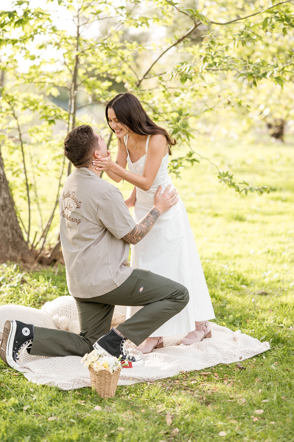 Woman in white dress smiling at kneeling man on picnic blanket in sunny park. Greenery surrounds them, with a small basket of flowers nearby.