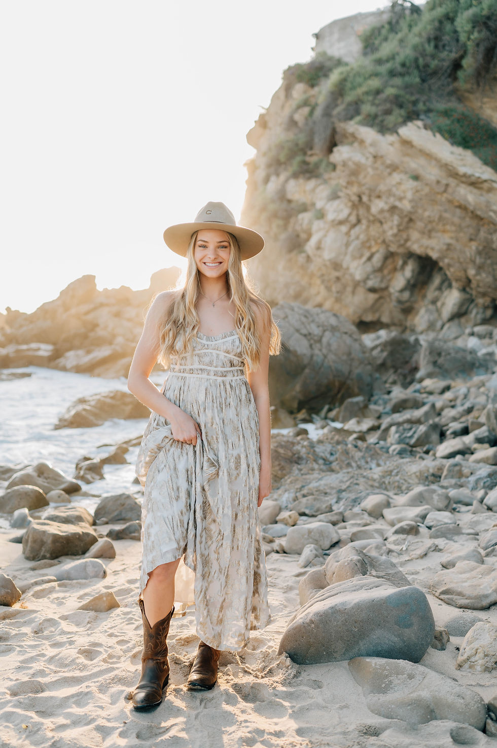 A girl in a flowing dress, cowboy boots, and a wide-brimmed hat stands on a rocky beach, smiling warmly as the sunlight casts a golden glow over the scene.