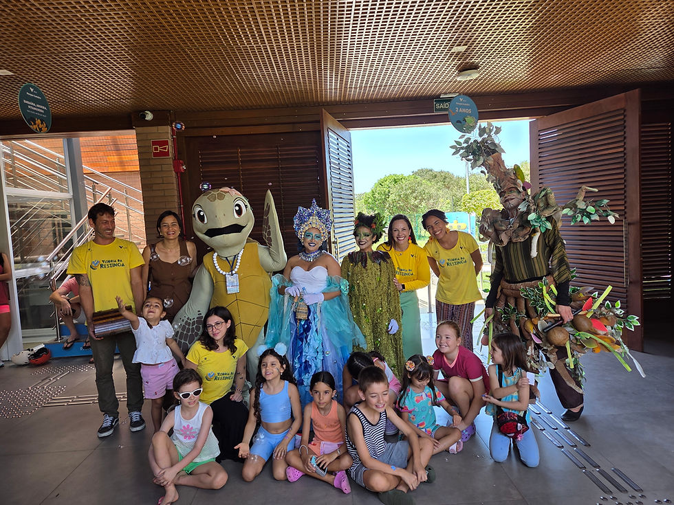 Grupo de crianças e adultos fantasiados posa sorridente em espaço coberto, com mascotes.