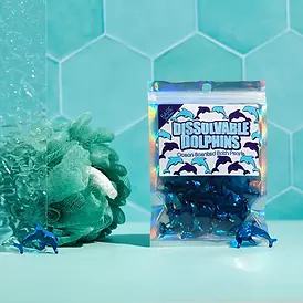Blue dolphin bath pearls in a resealable plastic packet standing next to a light green bath scrunchie