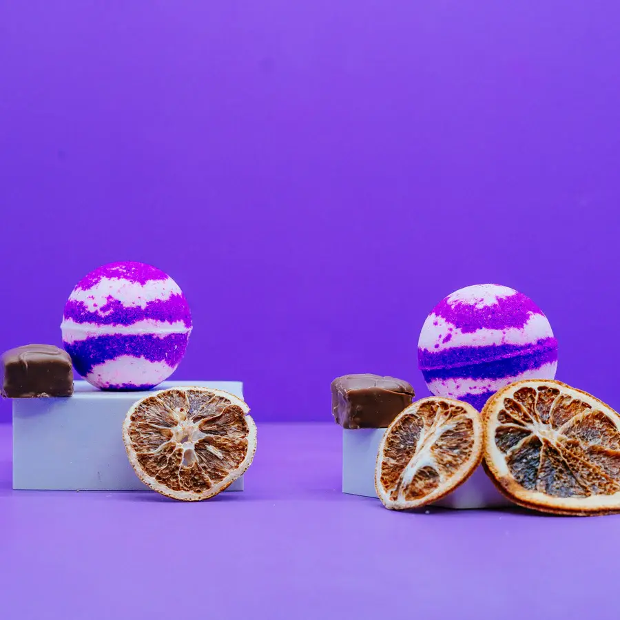 Two purple and white chocolate orange scented bath bomb on light purple plinth and with three dried oranges purple background