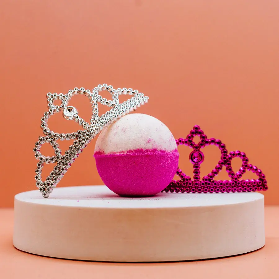 A pink and white ice queen bath bomb with two princess style tiaras on a white plinth with orange background