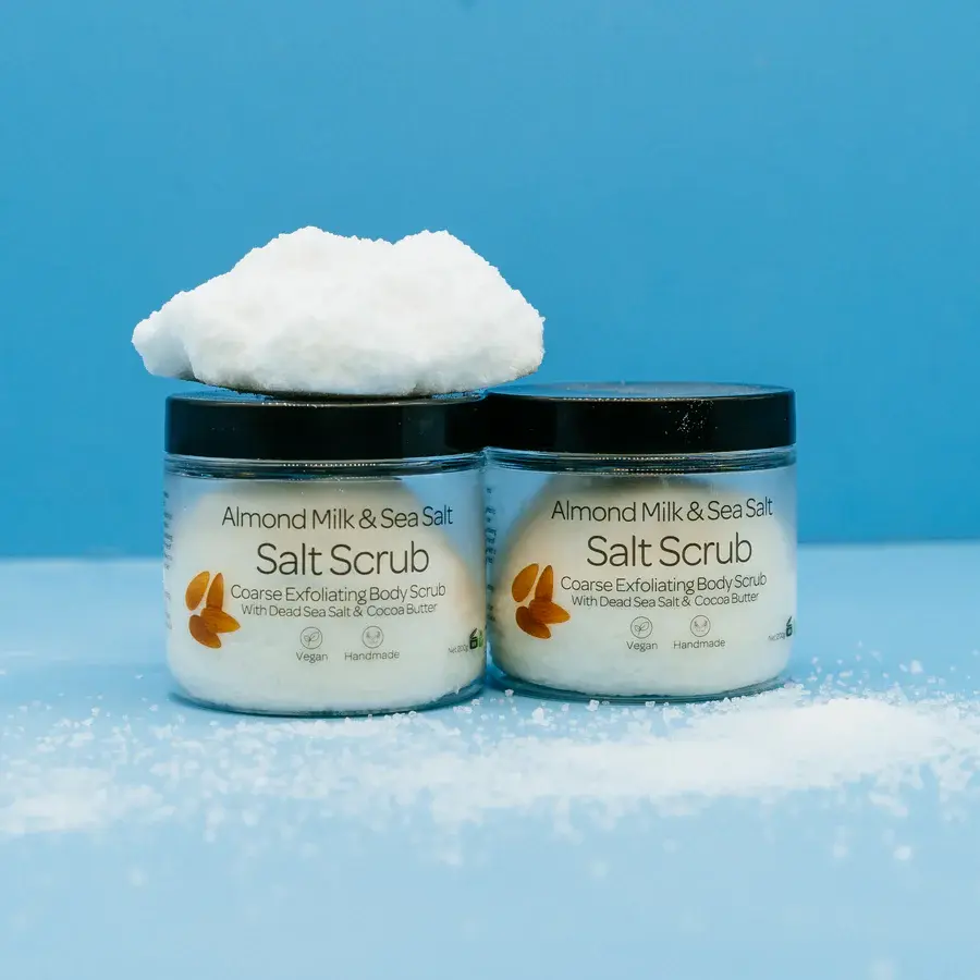 Two tubs of Almond Milk and Dead Sea Salt Scrub with a block of salt on top sprinkles of salt in front and blue background