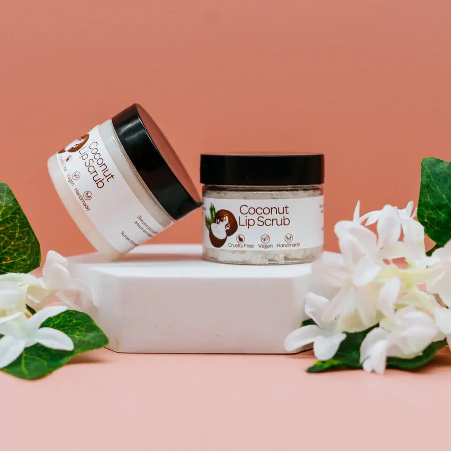 Two coconut lip scrub tubs one leaning against the other both on a white box with flowers with pink and orange background