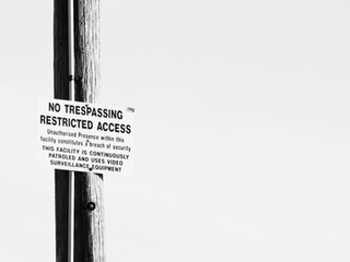 A white sign with black letters nailed onto a wooden pole writes: "NO TRESPASSING RESTRICTED ACCESS Unauthorized Presence within this facility constitutes a breach of security THIS FACILITY IS CONTINUOUSLY PATROLED AND USES VIDEO SURVEILLANCE EQUIPMENT"