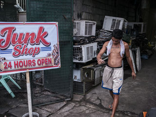 A shirtless man with a towel draping over his shoulders, wearing white board shorts and flip-flops, walks past dismantled air conditioners and a sign that writes, "Junk Shop 24 HOURS OPEN".
