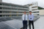 Two Hong Kong male teachers in business attire stand on top of a school roof, next to solar panels