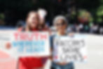 A white woman holds up a "TRUTH WHEREVER IT LEADS" sign. To her right, an elderly Asian woman holds a "Vaccines SAVE LIVES sign"