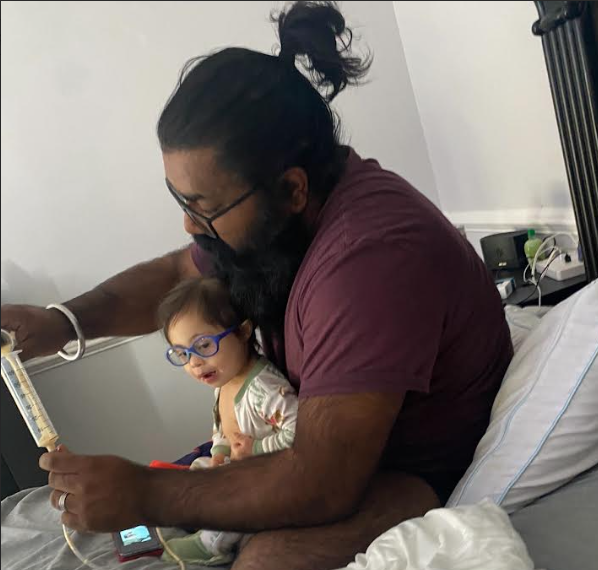 Jaswinder, a South Asian man, studies medication, as the baby on his lap looks on.
