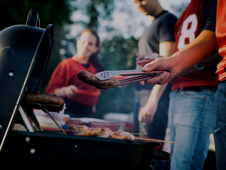 People grill sausages outdoors, one in a red jersey uses tongs beside a black barbecue at a smoky backyard cookout.