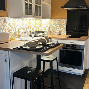 Cuisine moderne rénovée, meubles blancs, plan de travail bois clair, crédence motifs géométriques, hotte noire, îlot central avec tabourets noir et blanc, table dressée.