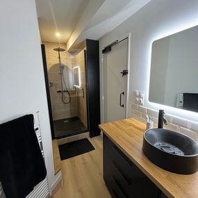 Salle de bain rénovée, douche paroi verre cadre noir, carrelage métro, meuble vasque noir bois clair, vasque noire, miroir éclairé.