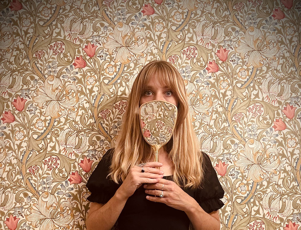 A blonde woman holds up a personal mirror in front of her nose and mouth and the back of the mirror has the same pattern as the wallpaper behind her.